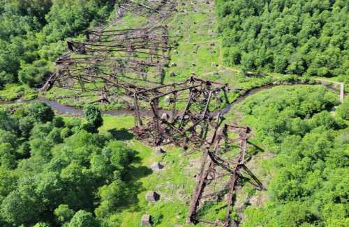 Aerial view of a collapsed metal structure surrounded by dense greenery and a winding stream.