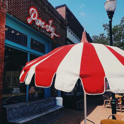 A red and white striped umbrella shades an ice cream shop with a vintage sign, set in a charming street scene.