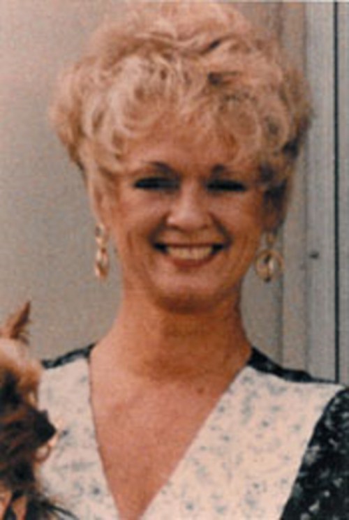 A smiling woman with curly blonde hair, wearing earrings, holding a small dog.