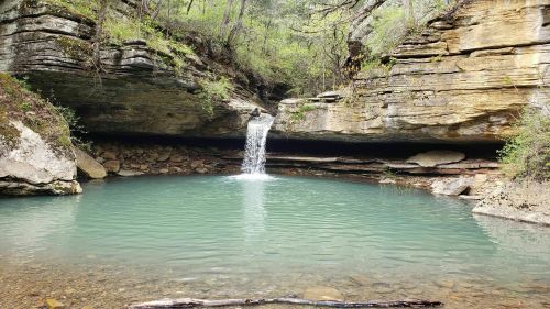 A serene natural scene featuring a small waterfall cascading into a turquoise pool surrounded by rocky cliffs and greenery.