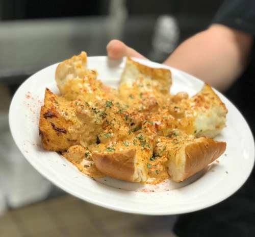 A person holds a plate of cheesy garlic bread topped with herbs and spices, with pieces cut and slightly toasted.