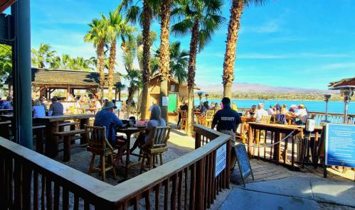 A lively outdoor bar scene with palm trees, tables, and patrons enjoying the view of a lake and mountains in the background.