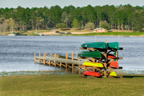 Colorful kayaks stacked on a rack by a wooden dock on a calm lake, surrounded by green trees and a distant cabin.