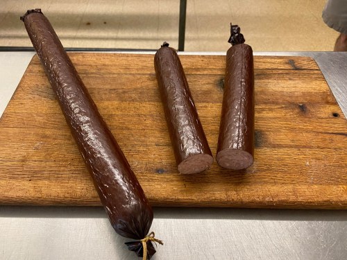 Three cylindrical sausages on a wooden cutting board, varying in size, with a shiny brown casing.
