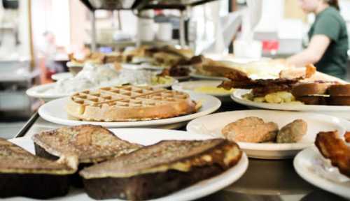 A variety of breakfast dishes on plates, including waffles, bacon, eggs, and toast, in a busy kitchen setting.