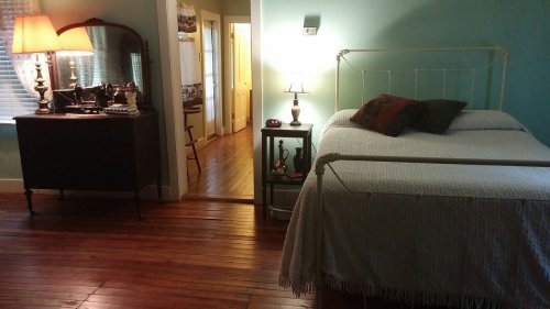 Cozy bedroom with a bed, vintage furniture, and warm lighting, featuring wooden floors and a doorway to another room.