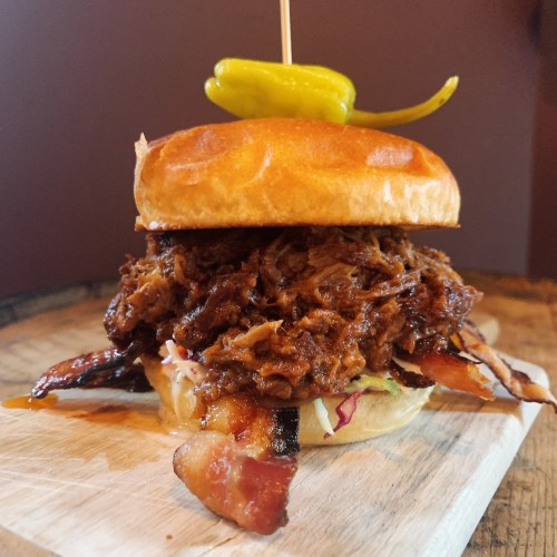 A hearty pulled pork burger topped with bacon and a pepper, served on a toasted bun with coleslaw.
