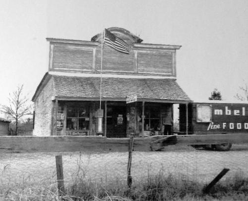 Black and white image of a vintage general store with an American flag, surrounded by a rural landscape.