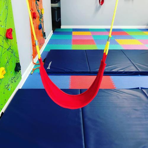 A colorful indoor play area featuring climbing walls, mats, and a red swing hanging from the ceiling.