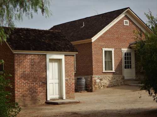 A brick house with a sloped roof and a small adjacent building, set on a dirt path surrounded by greenery.