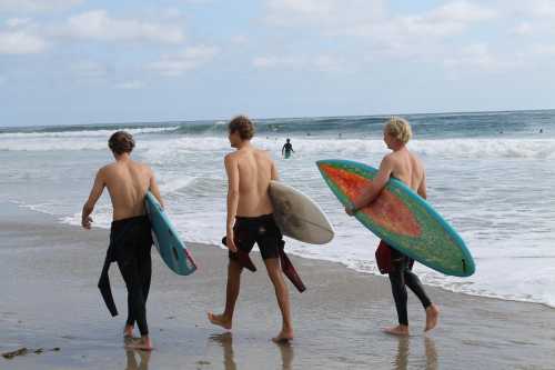 Three shirtless surfers walk along the beach, carrying their surfboards, with waves in the background.