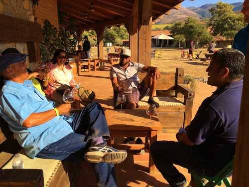 A group of four people relax outdoors, enjoying drinks and conversation under a wooden structure with mountains in the background.