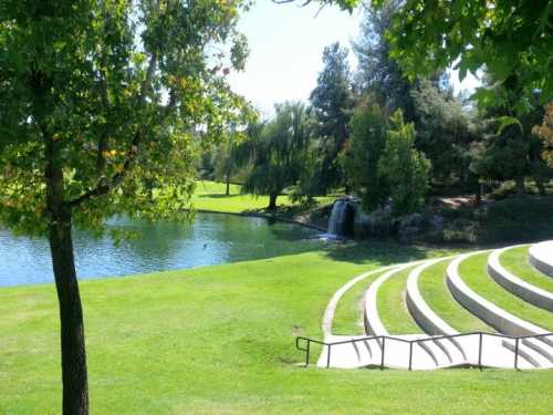 A serene park scene featuring a pond, waterfall, green grass, trees, and tiered seating.