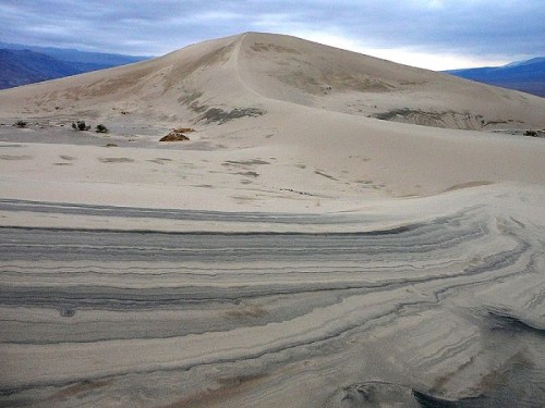 A vast desert landscape featuring a large sand dune and layered, textured ground under a cloudy sky.