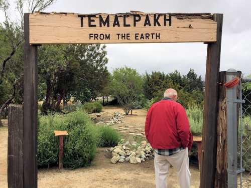 A person in a red jacket stands under a sign reading "TEMALPAKH FROM THE EARTH" in a natural outdoor setting.