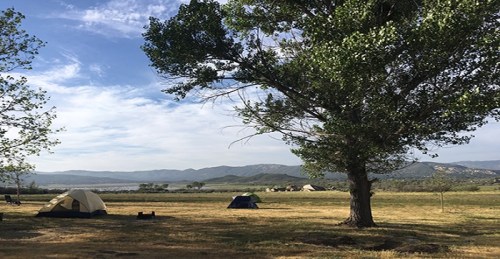 A serene campsite with tents under a large tree, set against a backdrop of mountains and a clear blue sky.