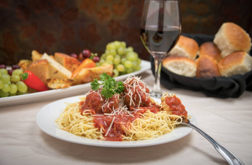 A plate of spaghetti with meatballs and marinara sauce, garnished with parsley, alongside a glass of red wine and bread rolls.