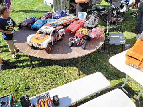 A table outdoors displays various remote-controlled cars, with people and chairs in the background.