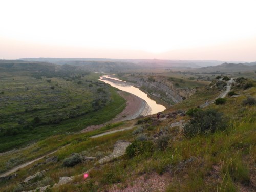 A winding river flows through a lush valley at sunset, surrounded by rolling hills and greenery.