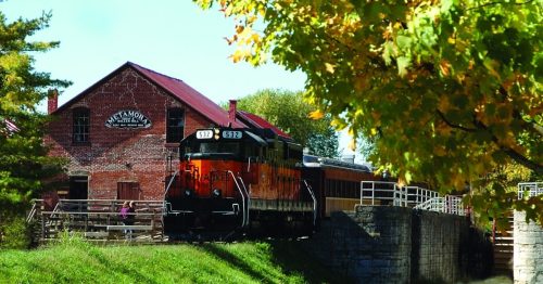 A vintage train near a red brick building by a calm waterway, surrounded by trees with autumn leaves.