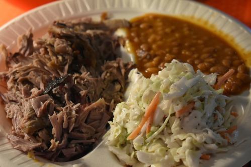 A plate with pulled pork, coleslaw, and baked beans, served on a disposable paper plate.