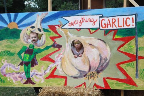 Two children peek through a colorful garlic-themed cutout, one dressed as a superhero, with a vibrant background.