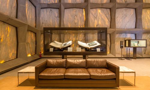 Modern interior featuring a brown leather sofa, a glass display case with open books, and textured stone walls.