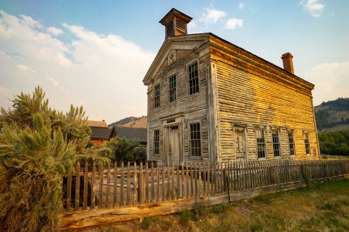 Historic wooden house with a peaked roof, surrounded by a wooden fence and overgrown grass, under a warm sunset sky.
