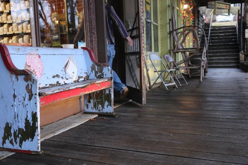 A weathered blue bench with peeling paint sits on a wooden walkway outside a shop, with a person exiting through the door.