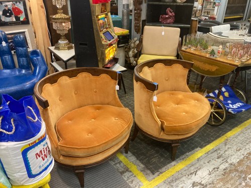 Two vintage, tufted orange chairs with wooden legs, surrounded by various eclectic items in a thrift store.