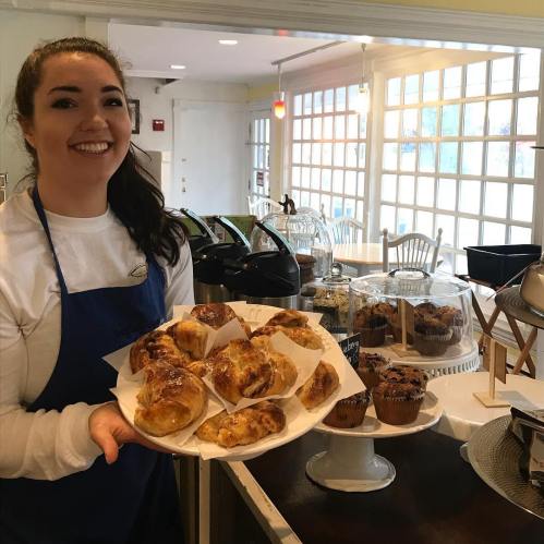 A smiling woman in an apron holds a plate of pastries in a cozy café setting.