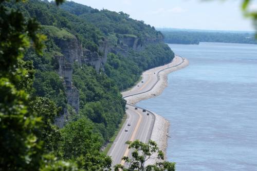 A winding road along a river, flanked by lush green cliffs and trees under a clear blue sky.