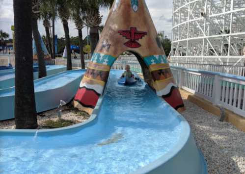 A child rides a water slide shaped like a teepee, surrounded by palm trees and a roller coaster in the background.