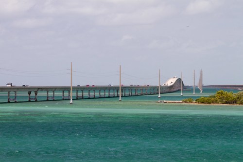 A long bridge stretches over turquoise waters, with cars traveling and sailboats in the distance under a cloudy sky.