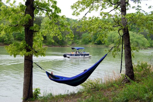 A blue hammock hangs between two trees by a calm lake, with a boat cruising in the background.