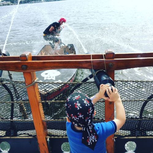 A child in a pirate hat watches a water-splashing boat on a lake, with a pirate flag visible on the boat.