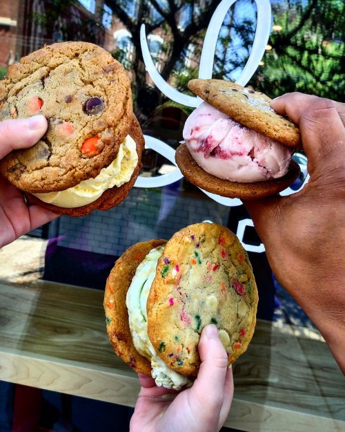 Three hands hold colorful cookie ice cream sandwiches, featuring various ice cream flavors, against a shop window backdrop.
