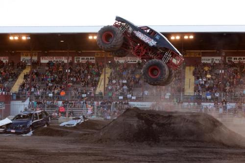 A monster truck leaps over a dirt ramp, with a crowd watching in the background at an outdoor event.