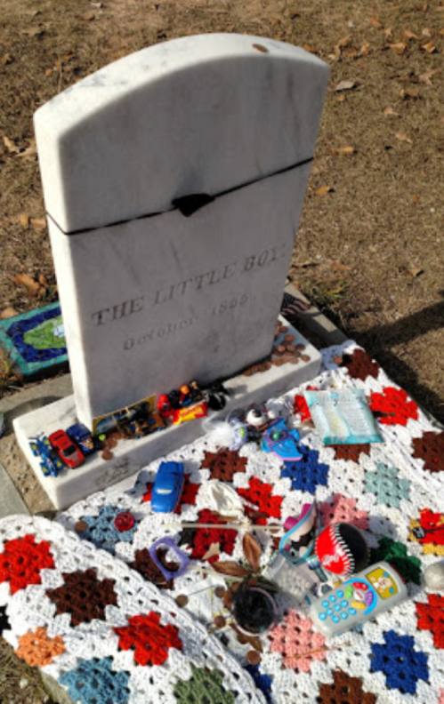 A gravestone reading "THE LITTLE BOY" surrounded by toys and a colorful crocheted blanket.