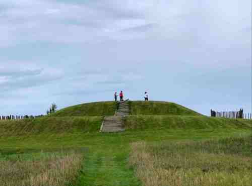 Three people stand on a grassy mound with steps, surrounded by open fields and a cloudy sky.
