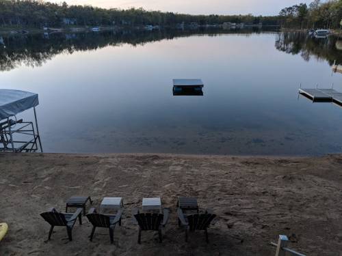 A serene lake view with empty chairs on the sandy shore and a floating dock in the distance.