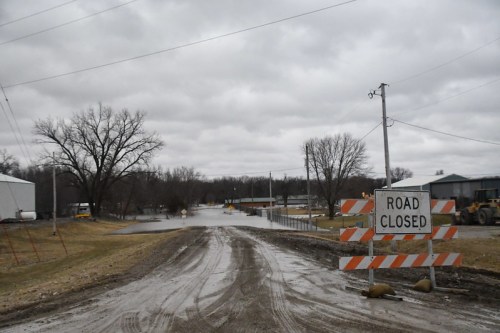 A flooded road with a "Road Closed" sign, surrounded by overcast skies and bare trees.