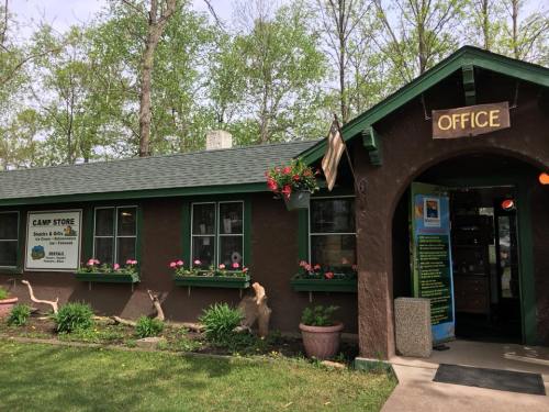 A rustic camp office with green trim, flower boxes, and a sign for the camp store, surrounded by trees.