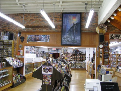 Interior of a comic book store with shelves of comics, a large Batman painting, and bright overhead lights.
