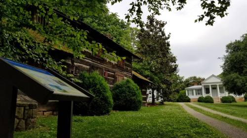 A rustic log cabin and a white house are surrounded by greenery, with a sign in the foreground on a gravel path.
