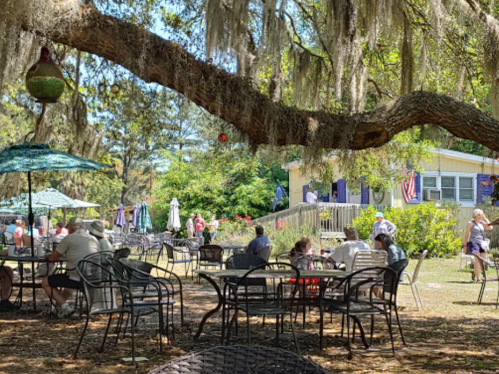 A sunny outdoor café scene with people dining under trees, featuring tables, umbrellas, and a building in the background.