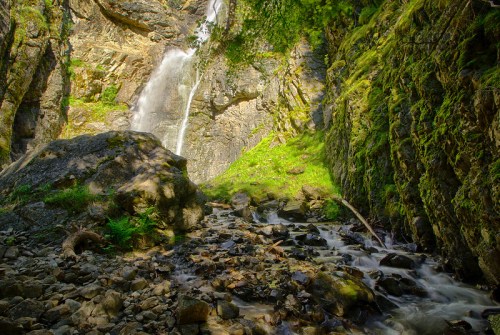 A serene waterfall cascades down rocky cliffs, surrounded by lush greenery and a flowing stream below.