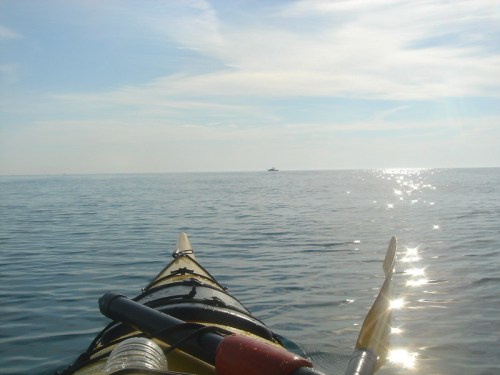 A kayak on calm water, reflecting sunlight, with a distant boat on the horizon under a clear blue sky.