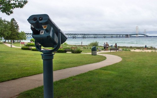 A scenic view of a park with a telescope, path, and a bridge in the background over a body of water.