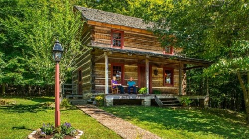 A rustic log cabin surrounded by trees, featuring a porch with two people sitting and a lamp post in the front yard.